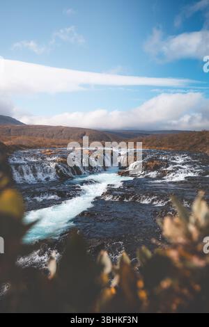 La cascata Bruarfoss in Islanda, conosciuta per le sue acque blu vivide e la roccia lavica scura, scorre attraverso un terreno colorato d'autunno sotto un cielo fresco. Una ge nascosta Foto Stock