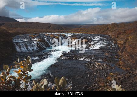 La cascata Bruarfoss in Islanda, conosciuta per le sue acque blu vivide e la roccia lavica scura, scorre attraverso un terreno colorato d'autunno sotto un cielo fresco. Una ge nascosta Foto Stock