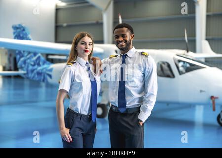 Due piloti di compagnie aeree sorridenti che si pongono con sicurezza di fronte a un jet privato all'interno di uno spazioso hangar, mostrando il loro cameratismo professionale Foto Stock