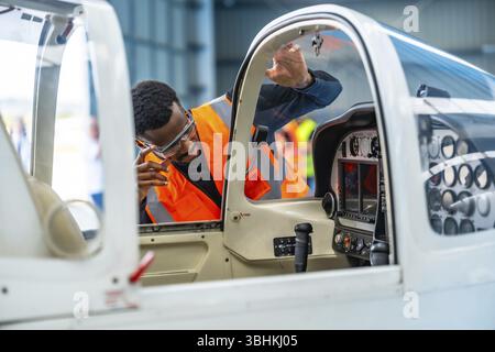 Un ingegnere aeronautico che indossa occhiali di sicurezza e un giubbotto ad alta visibilità ispeziona la cabina di pilotaggio di un piccolo aereo all'interno di un hangar Foto Stock