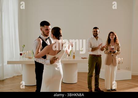 Giovane uomo adulto caucasico che balla con una giovane donna adulta caucasica in abito da sposa mentre uomo e donna in piedi sullo sfondo applaudono Foto Stock