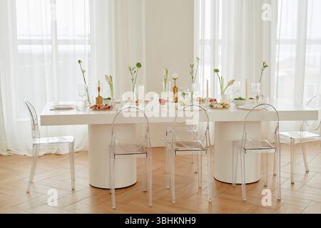 Tavolo vuoto per la celebrazione di nozze con piatti, bicchieri, candele e composizioni floreali in piedi in una sala luminosa, sedie trasparenti intorno al tavolo, nessuna persona visibile Foto Stock