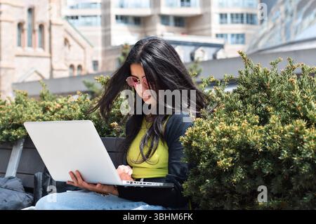 Giovane donna seduta sulla panchina fuori usando un computer portatile Foto Stock