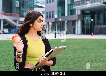 Giovane studentessa che cammina nel campus con un computer portatile e una tazza di caffè Foto Stock