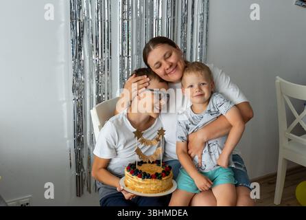 Famiglia felice che festeggia il compleanno a casa con una torta fatta in casa decorata con frutti di bosco e topper stellato d'oro. Madre che abbraccia due bambini con l'argento Foto Stock