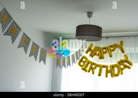 Happy Birthday palloncini a forma di lamina d'oro e decorazioni colorate per feste appese in un luminoso soggiorno moderno con bandiere a grappolo. Casa festiva Foto Stock
