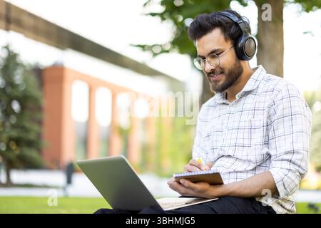 Un giovane uomo che lavora su un notebook all'aperto, indossa le cuffie e scrive su un notebook. Lo sfondo mostra alberi e un edificio. Foto Stock