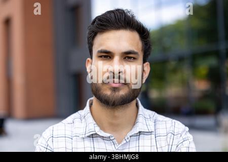 Un uomo indiano mirato guarda direttamente alla fotocamera, scattato in primo piano. Sta indossando una camicia a scacchi fuori. Foto Stock