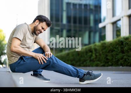 Un giovane uomo stringe il ginocchio, probabilmente avvertendo dolore durante o dopo una sessione di allenamento all'aperto. Foto Stock