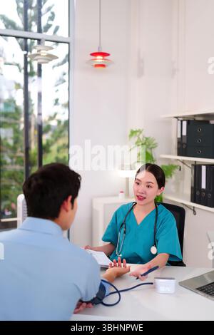 Una donna asiatica medico controlla la pressione sanguigna di un maschio adulto durante una visita medica. Discutono di salute in un ambiente clinico moderno per Foto Stock