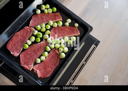 Bistecche di maiale crude e cavoletti di Bruxelles su vassoio del forno pronti per la tostatura in cucina, attenzione selettiva Foto Stock