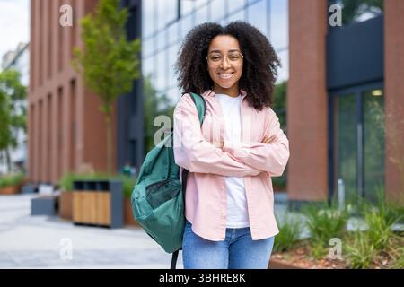 Una giovane donna afroamericana sorridente che indossa occhiali e uno zaino si trova di fronte a un edificio moderno. Foto Stock