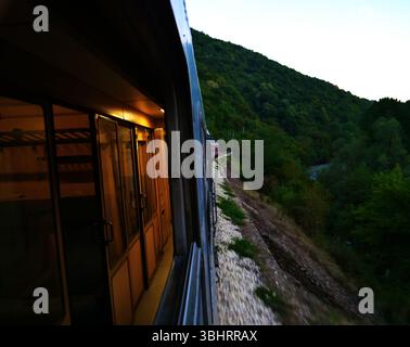 Treno bulgaro sulla strada da Bucarest a Sofia passando attraverso la foresta panoramica e le colline durante il crepuscolo Foto Stock