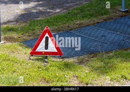 Un cartello triangolare rosso e bianco si trova in erba verde accanto a una griglia metallica. Sullo sfondo viene visualizzata un'area pavimentata grigia. Foto Stock