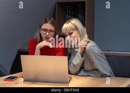 Due donne d'affari professionali nell'ufficio dell'azienda vinicola che collaborano alla strategia di marketing, incentrata sullo schermo di un notebook, circondate da energia positiva, branding Foto Stock
