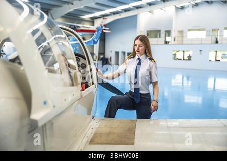 Pilota donna sicura di sé che sale a bordo di un piccolo aereo in un hangar, preparandosi per un volo emozionante e abbracciando l'avventura che ti attende Foto Stock