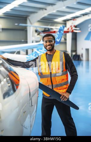 Sorridente ingegnere afroamericano dell'aviazione che tiene in mano una pala elica, esegue controlli di manutenzione in un hangar di aeromobili Foto Stock