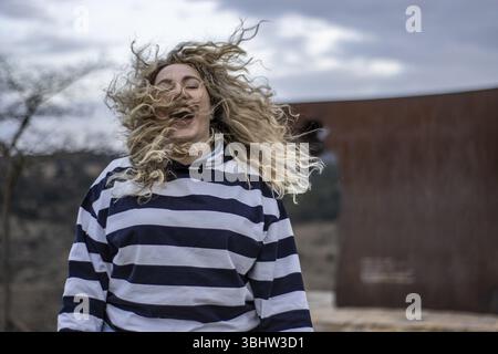 Vista frontale di una donna che ride con gli occhi chiusi e i capelli che soffiano al vento all'aperto indossando un indumento a righe Foto Stock