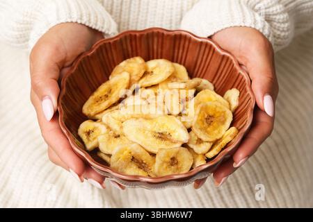 Gocce di banana essiccate in un recipiente di ceramica. Mani che tengono uno spuntino dolce. Frutta tropicale croccante. Sana alternativa alle caramelle. Consistenza naturale degli alimenti secchi. Foto Stock