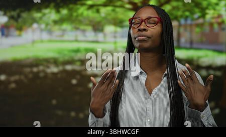 Giovane donna con capelli ricci nel parco che indossa occhiali caldi sotto il sole in un ambiente urbano all'aperto. Foto Stock