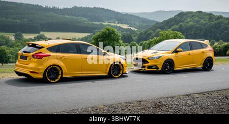 Due Focus STS color mandarino parcheggiate su un lato della strada rurale. Vista laterale di due Ford berlini nel paesaggio rurale delle montagne Eifel. Foto Stock