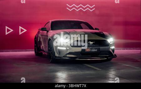 Ford Mustang beige camo nel garage. Vista frontale di tre quarti del pony americano parcheggiato vicino al muro color marrone illuminato. Foto Stock