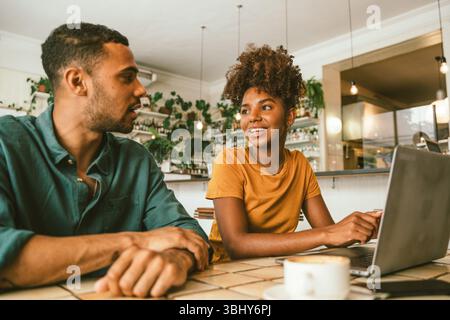 Giovane donna che usa un computer portatile e parla con un uomo seduto insieme in un bar Foto Stock