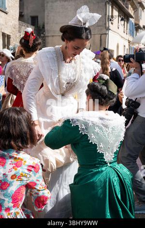 La regina regnante in bianco si erge fuori dalla chiesa, mentre due donne adattano il suo lungo vestito all'inizio del Festival del costume dove oltre 50 persone Foto Stock