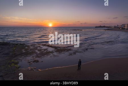 Una donna si erge sulla spiaggia sabbiosa, affascinata dall'accattivante tramonto sopra le calme acque di Punta secca, Sicilia, Italia Foto Stock