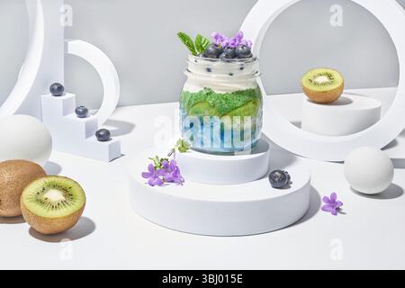 Un vivace parfait di yogurt in un barattolo con fette di kiwi fresche, mirtilli e artistiche tonalità verde e viola, presentato su uno sfondo bianco minimalista con wi-fi Foto Stock
