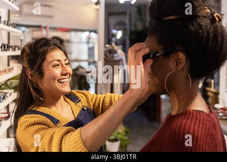 Un allegro assistente di negozio aiuta un cliente a provare un paio di eleganti occhiali da sole in un'accogliente boutique. Gli scaffali pieni di bicchieri creano un negozio accogliente Foto Stock