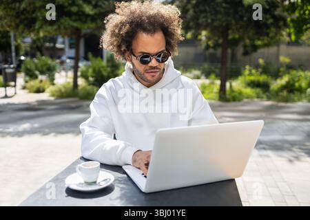 Un uomo d'affari con acconciature afro, con occhiali da sole e una felpa bianca, lavora su un computer portatile in un bar all'aperto. Una tazza di caffè si trova nelle vicinanze, con Foto Stock