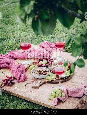 Un affascinante set da picnic con bicchieri di rosa, ciotole di cracker croccanti e uva verde e rossa fresca, elegantemente sistemati su una tavola di legno, con l'ingegno Foto Stock