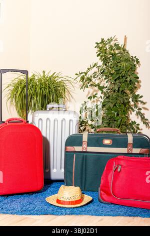 Una collezione di vivaci valigie da viaggio, tra cui il rosso e l'argento, è disposta accanto a lussureggianti piante verdi, con un cappello di paglia adagiato su un tappeto blu in un laccio Foto Stock