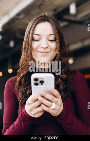 Una giovane studentessa con i capelli lunghi sorride mentre guarda il suo smartphone, indossando un abito marrone. È al chiuso e sembra che si stia divertendo Foto Stock
