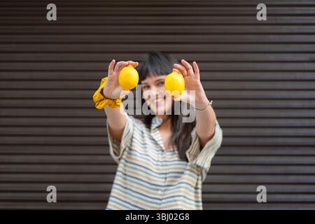 Una giovane donna sorridente tiene due limoni gialli vibranti verso la fotocamera, indossando una camicia a righe e stando in piedi di fronte a uno sfondo di legno scuro, c Foto Stock