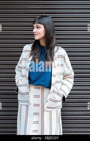 Una donna si staglia con sicurezza su uno sfondo in metallo ondulato e indossa un elegante outfit a righe. La sua posa rilassata e lo sfondo neutro creano un Foto Stock