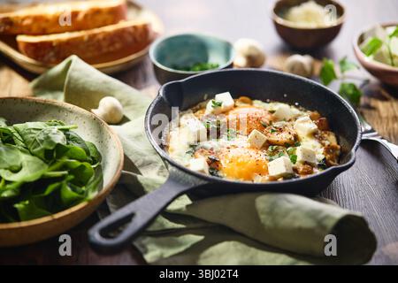 Uova fritte con spinaci, funghi, formaggio grattugiato e formaggio feta in piccola padella di ghisa Foto Stock
