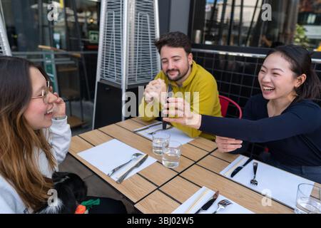 Un gruppo eterogeneo di amici condivide risate e cucina asiatica in un ristorante all'aperto. Un cane si siede in grembo di un amico, creando un caldo e gioioso scen Foto Stock