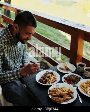 Uomo che si gode un pranzo tradizionale balcanico all'aperto, seduto accanto a un tavolo in legno con terrazza pieno di cibo. Foto Stock