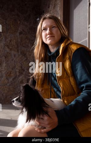 Una persona in un gilet giallo siede con il suo cane papillon di razza pura in grembo, catturando un momento sereno alla luce del sole. La scena trasmette calore e. Foto Stock