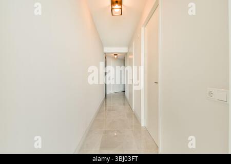 Un corridoio luminoso e minimalista con pareti e porte bianche, caratterizzato da piastrelle eleganti e moderne luci a sospensione che emettono un caldo bagliore, creando un invitante bancomat Foto Stock