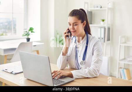 Medico seduto sul posto di lavoro che parla con il telefono cellulare e che lavora con il computer portatile in clinica. Foto Stock