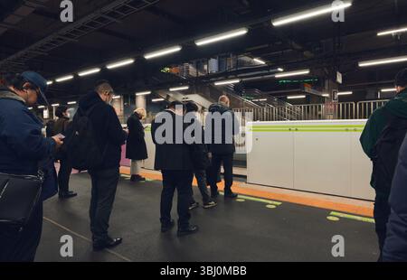 I pendolari attendono in linee ordinate su una piattaforma ferroviaria di Tokyo sotto il fresco bagliore delle luci fluorescenti Foto Stock
