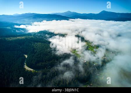 Affascinante vista aerea del paesaggio ricoperto da soffici nuvole bianche. Lussureggianti vallate verdi e tortuose strade sbirciano, mentre le lontane montagne blu si stagliano maestosamente sotto un cielo chiaro e limpido. Foto Stock