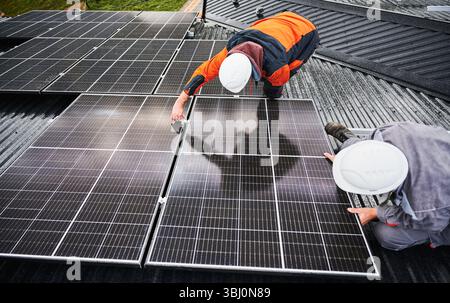 Tecnici professionisti che regolano i pannelli fotovoltaici. Installazione di impianti solari sul tetto della casa. Vista dall'alto di due lavoratori maschi in muri e tute da lavoro che fissano la batteria solare. Foto Stock