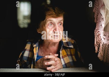 Una donna anziana siede tranquillamente, le mani piegate, guardando con attenzione verso la finestra. Il suo volto è calmo e concentrato Foto Stock