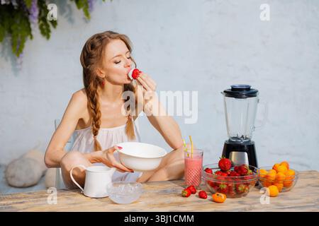 Donna che mangia fragole con panna montata al tavolo all'aperto con frullato, frullatore e ciotole di frutta fresca. Atmosfera da dessert estiva. Foto Stock