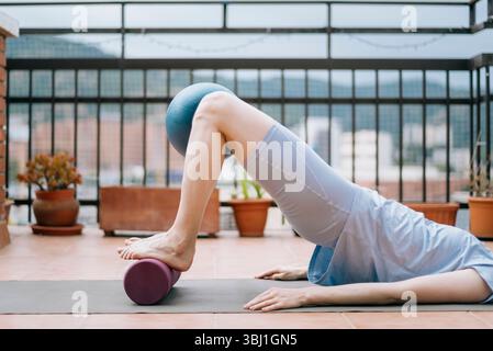 Donna che fa glute bridge con rullo in schiuma e palla Pilates per l'allenamento in ipermobilità Foto Stock