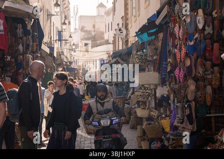 Essaouira, Marocco - 23 febbraio 2025: Un uomo locale guida uno scooter nelle strade affollate della medina di Essaouira, fiancheggiata da negozi di souvenir. Foto Stock